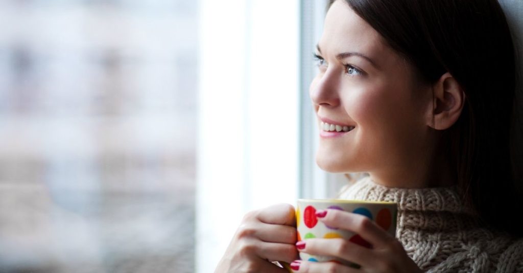 woman looking out of the window and smiling whilst holding a mug